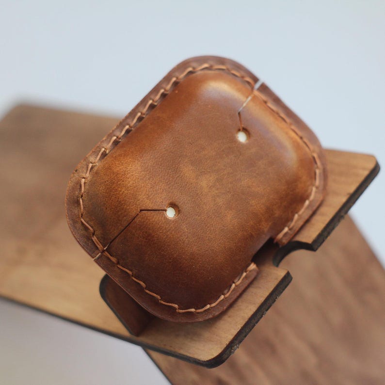 May include: A brown leather case with stitching around the edges. The case has two small holes in the top. The case is sitting on a wooden stand.