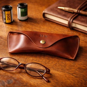 Könnte beinhalten: Ein braunes Lederetui für Brillen mit den Initialen "MW" auf der Vorderseite, gesichert durch einen goldfarbenen Druckknopf. Das Etui befindet sich neben einer Brille und einem ledergebundenen Notizbuch. Zwei Filmrollen sind ebenfalls sichtbar.