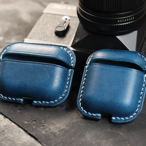 May include: Two blue leather cases for Airpods with light blue stitching. The cases are sitting on a wooden surface.