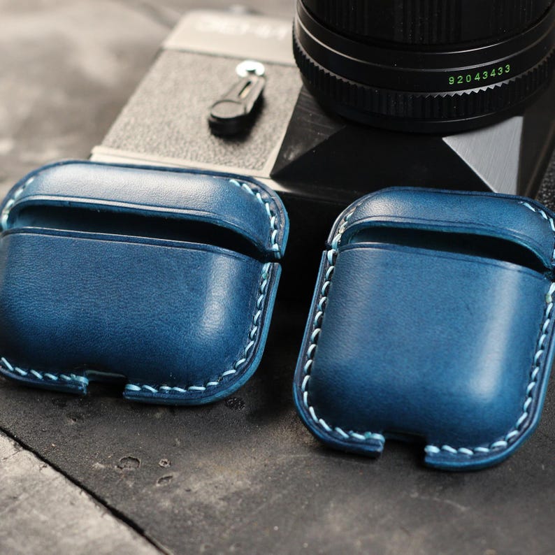 May include: Two blue leather cases for Airpods with light blue stitching. The cases are sitting on a wooden surface.