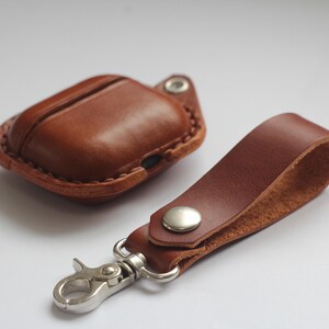 May include: A brown leather case for AirPods with a matching brown leather strap and a silver metal carabiner clip.