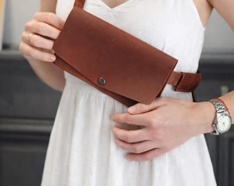 May include: A brown leather fanny pack with a flap closure and a strap. The bag is worn across the body. The fanny pack has a button closure. The strap is brown. The person is wearing a white dress.