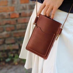 Op de afbeelding: Bruin leren crossbody telefoontasje met een voorvak en een lange, dunne riem. De tas is rechthoekig met afgeronde hoeken. Achtergrond: bakstenen muur.