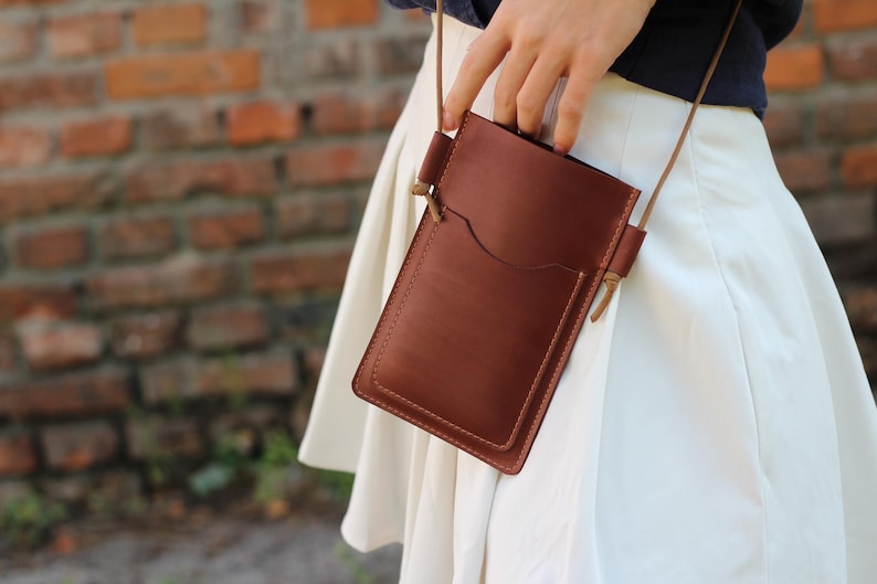 Op de afbeelding: Bruin leren crossbody telefoontasje met een voorvak en een lange, dunne riem. De tas is rechthoekig met afgeronde hoeken. Achtergrond: bakstenen muur.