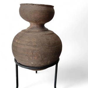 May include: A brown, textured ceramic vessel with a bulbous base and a smaller, bowl-shaped top. The vessel is supported by a black metal stand. The ceramic piece has horizontal and vertical etched lines, giving it a rustic appearance. The overall design is reminiscent of ancient pottery.