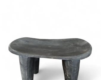 May include: A small, low-profile stool carved from a single piece of dark, charcoal-colored wood. The seat is rectangular with rounded edges and a slightly concave surface, supported by four thick, tapered legs with visible chisel marks.