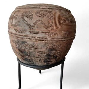 May include: A large, round, earthenware vessel with intricate, raised patterns and designs. The vessel is a warm, earthy brown colour and sits on a black metal stand. The patterns include dots, lines, and geometric shapes, suggesting an ancient or tribal aesthetic.