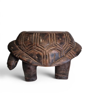May include: A dark brown, wooden turtle-shaped stool. The stool has a carved shell design and four short legs. The head and tail of the turtle are also carved. The stool is a decorative piece of furniture.