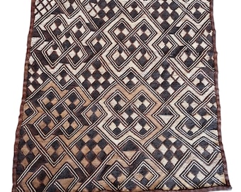 May include: A square textile piece featuring a repeating geometric pattern in shades of dark brown, tan, and cream. The design consists of interlocking diamond shapes and knotted lines, finished with a solid dark brown border on all four sides.