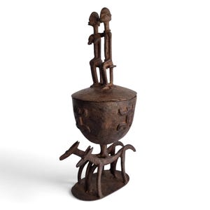 May include: A dark brown metal sculpture featuring a bowl-shaped container with a lid. Two stylized figures sit on the lid, while the base is supported by four animal figures standing on an oval platform. The bowl has embossed symbols.