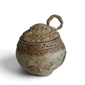 May include: A round, light green ceramic jar with a textured, weathered finish. The lid features a braided handle and embossed wavy line patterns. The body of the jar displays a similar wavy pattern around the top and a spiral design.