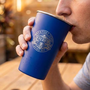 May include: A blue stainless steel pint glass with a silver rim and a logo that reads "BREWHAT TERBY RHERIFIR BEWH DFC". The glass is being held by a person and is being used to drink from.
