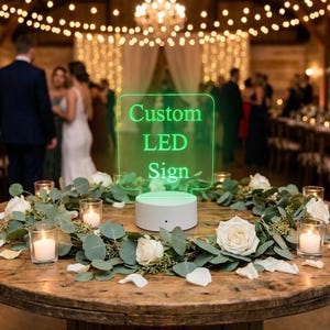May include: A rectangular clear acrylic sign with green LED illumination displaying the text Custom LED Sign, mounted on a white circular base, positioned in the center of a round wooden table decorated with a eucalyptus and white rose garland.