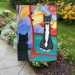 May include: A garden flag featuring a whimsical painting of two cats. One cat is black, facing a mirror, while the other is a tuxedo cat with a white chest and paws. The background has colorful abstract shapes.