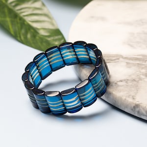 Könnte beinhalten: Ein blaues und weiß gestreiftes Armband aus rechteckigen Perlen. Das Armband liegt auf einer weißen Marmorfläche.