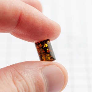 May include: A small, rectangular amber bead with a polished surface. The bead is a translucent, warm brown color with flecks of gold and darker brown inclusions. The bead is held between two fingers against a white background.