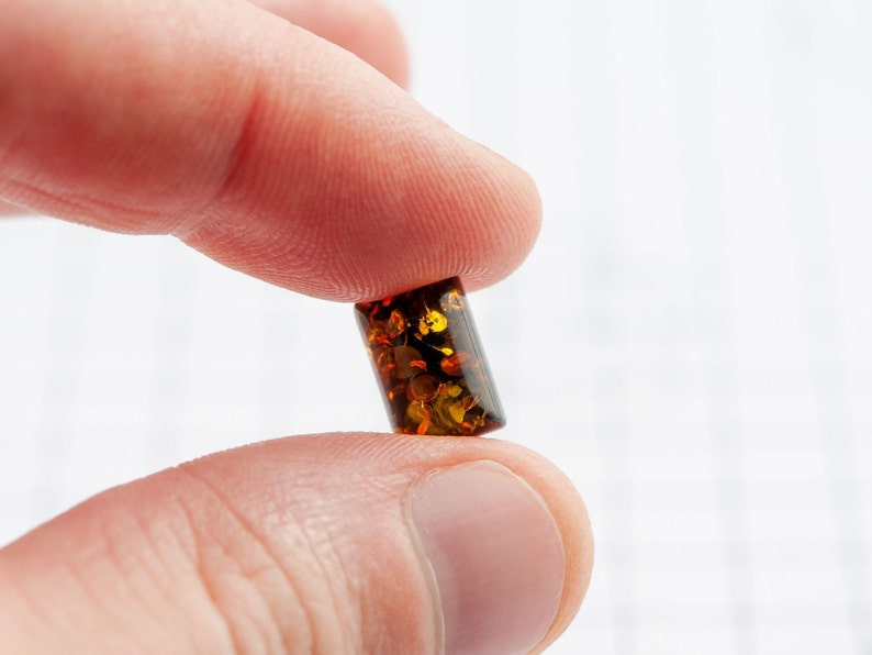 May include: A small, rectangular amber bead with a polished surface. The bead is a translucent, warm brown color with flecks of gold and darker brown inclusions. The bead is held between two fingers against a white background.