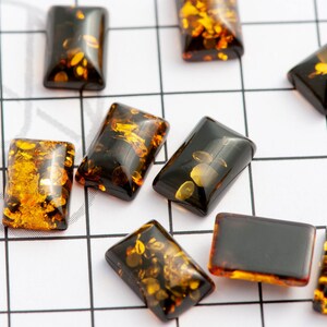 May include: Rectangular amber-colored beads with dark brown and golden inclusions are scattered on a white grid. The beads have a polished, glossy finish, and vary slightly in the distribution of the golden flecks within the translucent amber.