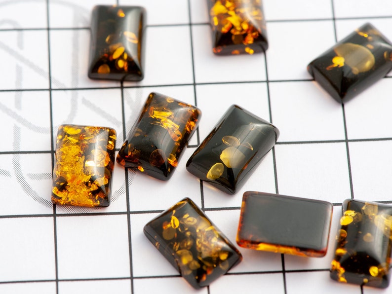 May include: Rectangular amber-colored beads with dark brown and golden inclusions are scattered on a white grid. The beads have a polished, glossy finish, and vary slightly in the distribution of the golden flecks within the translucent amber.
