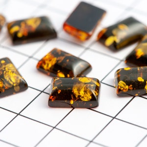 May include: Rectangular amber-colored stones with dark brown inclusions are scattered across a white grid. The stones vary in transparency, with some appearing nearly opaque and others showing intricate patterns of golden hues. The overall effect is a display of natural beauty.