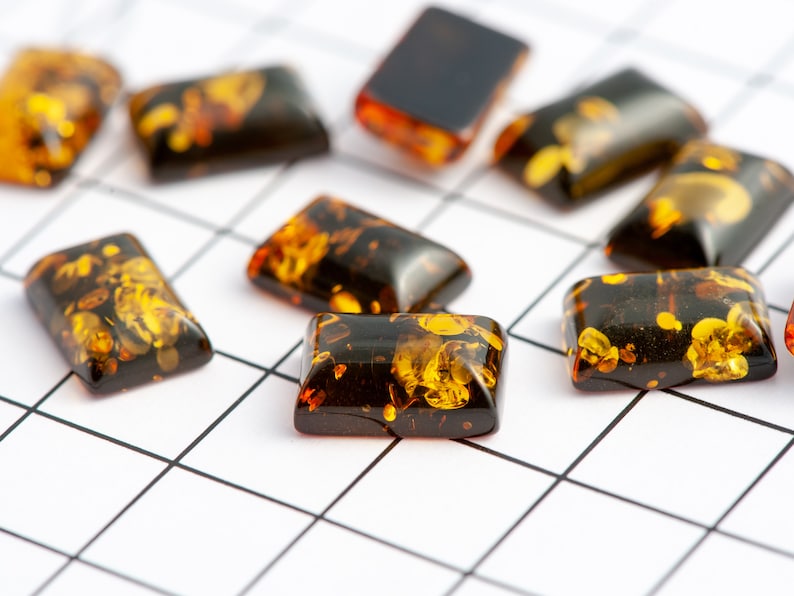 May include: Rectangular amber-colored stones with dark brown inclusions are scattered across a white grid. The stones vary in transparency, with some appearing nearly opaque and others showing intricate patterns of golden hues. The overall effect is a display of natural beauty.