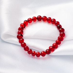 Può includere: Un braccialetto rosso di perline rotonde e lucidate. Il braccialetto è su uno sfondo di raso bianco.