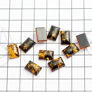 May include: A collection of rectangular amber-colored resin cabochons with inclusions of gold flecks. The cabochons are arranged on a white surface with a black grid pattern. The resin pieces vary in color from dark brown to golden yellow.