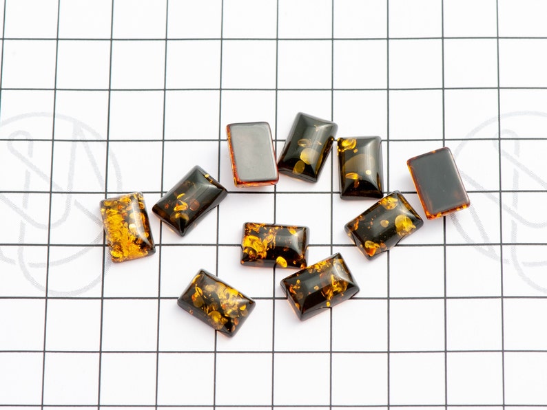 May include: A collection of rectangular amber-colored resin cabochons with inclusions of gold flecks. The cabochons are arranged on a white surface with a black grid pattern. The resin pieces vary in color from dark brown to golden yellow.