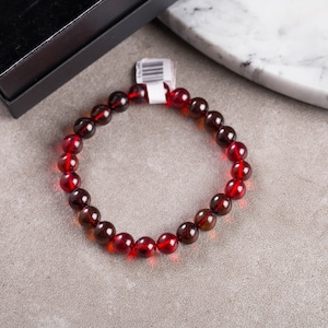 Może przedstawiać: Czerwona bransoletka z koralików z dołączoną metką, leżąca na szarej powierzchni. Bransoletka składa się z okrągłych, przezroczystych koralików w odcieniach czerwieni i brązu. Częściowo widoczne czarne pudełko.
