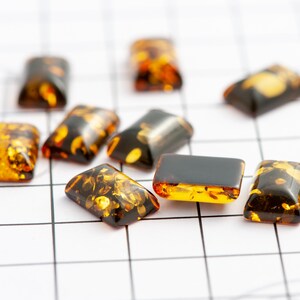 May include: A collection of rectangular amber-colored gemstones with dark brown and golden inclusions. The stones are arranged on a white grid, showcasing their polished surfaces and unique patterns. These gemstones are ideal for jewelry making.
