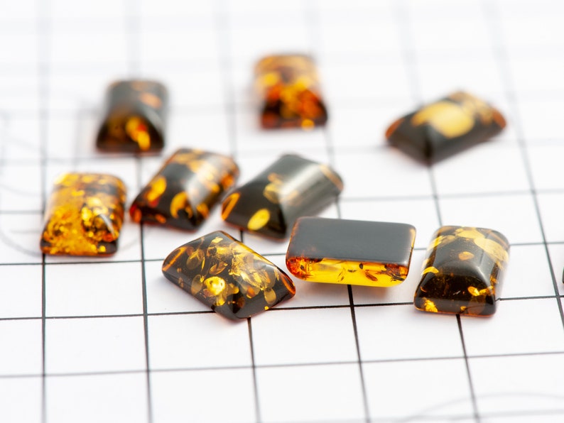May include: A collection of rectangular amber-colored gemstones with dark brown and golden inclusions. The stones are arranged on a white grid, showcasing their polished surfaces and unique patterns. These gemstones are ideal for jewelry making.