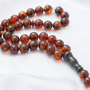 Puede incluir: Un rosario de cuentas de oración de color ámbar con una pieza final ornamentada de color marrón oscuro. Las cuentas son redondas y tienen un acabado brillante. El fondo es una tela blanca y sedosa.