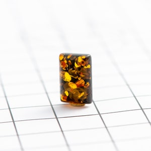 May include: A rectangular amber bead with a dark brown base and a mix of golden and brown inclusions. The bead is set against a white grid background, showcasing its translucent quality and natural patterns.
