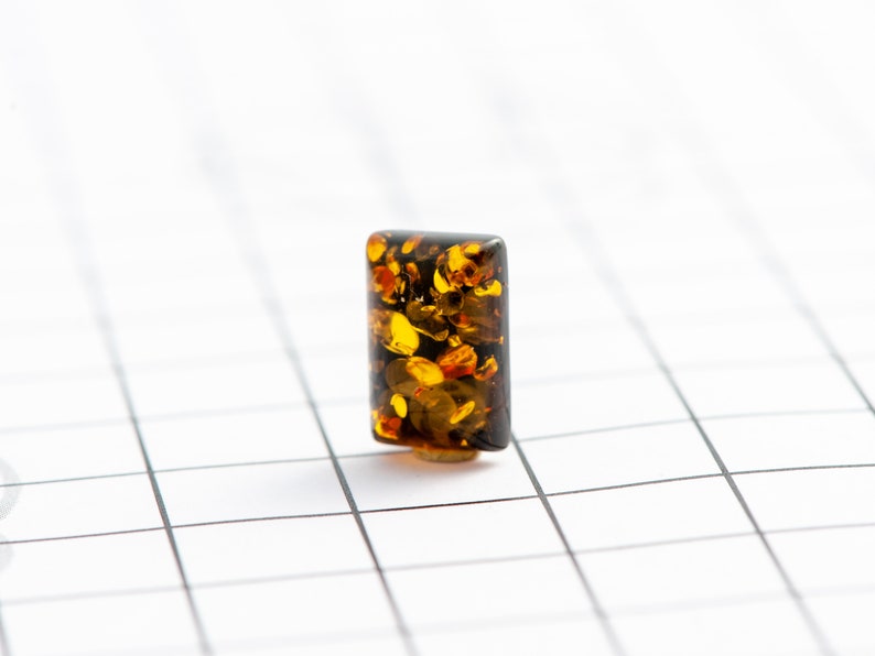 May include: A rectangular amber bead with a dark brown base and a mix of golden and brown inclusions. The bead is set against a white grid background, showcasing its translucent quality and natural patterns.