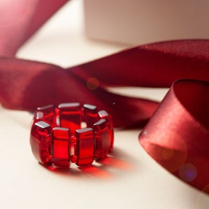 Op de afbeelding: Een rode glazen ring gemaakt van rechthoekige stukken. De ring ligt op een wit oppervlak met een rood satijnen lint op de achtergrond.