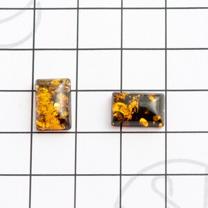 May include: Two rectangular amber cabochons with a mix of golden and dark brown hues. The cabochons are set against a white grid background, showcasing their polished surfaces and internal patterns. These gemstones are ideal for jewelry making.