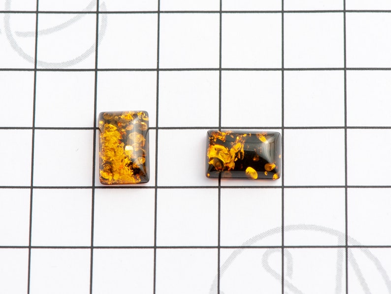 May include: Two rectangular amber cabochons with a mix of golden and dark brown hues. The cabochons are set against a white grid background, showcasing their polished surfaces and internal patterns. These gemstones are ideal for jewelry making.