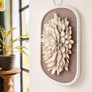 May include: A wall-mounted oval art piece featuring a white beaded frame and a taupe background. The center is adorned with a dense cluster of layered, white, petal-like shapes with gold-painted edges. The text 20 x 11 is printed at the bottom.