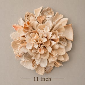 May include: A circular wall art piece composed of various light beige and cream-colored seashells and coral fragments arranged in a floral pattern. The arrangement measures 11 inches in diameter, featuring textured, spiraled, and flat shells.