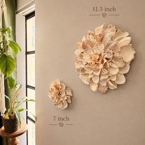 Op de afbeelding: Twee ronde wanddecoraties bestaande uit gelaagde, lichtgekleurde schelpen in een bloemvorm. Het grotere stuk heeft een diameter van 29,2 cm en het kleinere stuk heeft een diameter van 17,8 cm, beide bevestigd aan een muur.