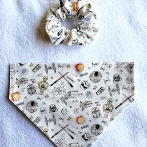 May include: White fabric scrunchie with a black and white Star Wars themed print. The scrunchie is paired with a white fabric bandana with a similar print. The bandana has a pointed bottom and is decorated with black and white drawings of Star Wars characters and objects, including Darth Vader, R2-D2, C-3PO, and the Millennium Falcon. The bandana also includes text such as "Rebel", "Rule the Galaxy", "Boom!" and "Kissesshh!"