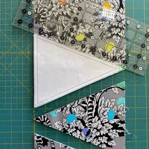 May include: A quilting project in progress, featuring patterned fabric with a black and white lemur and foliage design. Clear rulers are positioned over the fabric and a paper template, with cut fabric triangles visible. The fabric has colorful polka dots.
