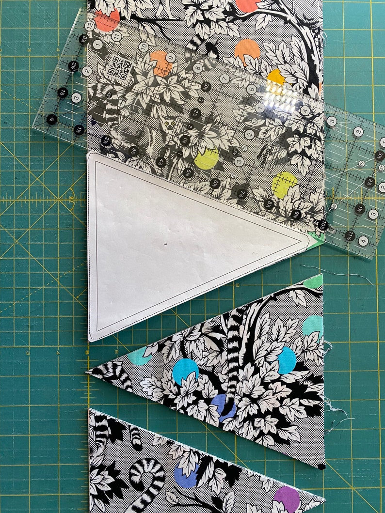May include: A quilting project in progress, featuring patterned fabric with a black and white lemur and foliage design. Clear rulers are positioned over the fabric and a paper template, with cut fabric triangles visible. The fabric has colorful polka dots.