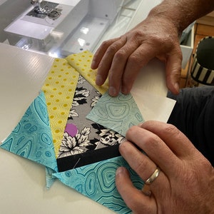 May include: A patchwork quilt block in progress, featuring various patterned fabrics in shades of blue, yellow, black, and white. The block is being held by hands, with a sewing machine visible in the background.
