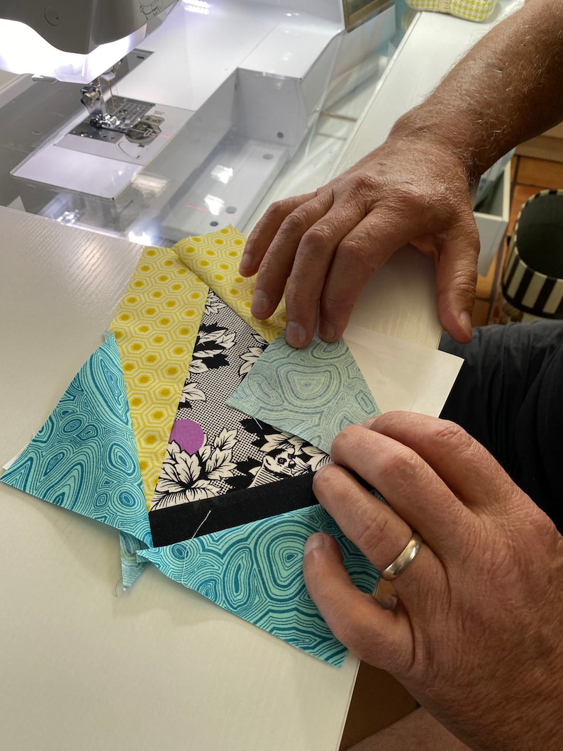 May include: A patchwork quilt block in progress, featuring various patterned fabrics in shades of blue, yellow, black, and white. The block is being held by hands, with a sewing machine visible in the background.