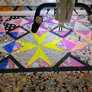 May include: A colorful quilt in progress, featuring geometric star patterns and various fabric designs. The quilt incorporates black, white, gray, pink, purple, blue, and yellow fabrics. A quilting machine is positioned above the quilt, with its needle poised to stitch.