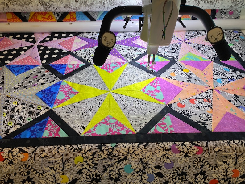 May include: A colorful quilt in progress, featuring geometric star patterns and various fabric designs. The quilt incorporates black, white, gray, pink, purple, blue, and yellow fabrics. A quilting machine is positioned above the quilt, with its needle poised to stitch.