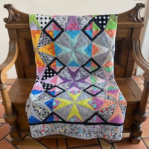 May include: A colorful patchwork quilt with star and diamond patterns, draped over a wooden bench. The quilt features various fabrics in shades of gray, black, yellow, orange, pink, purple, and blue. The quilt's border has a floral design.