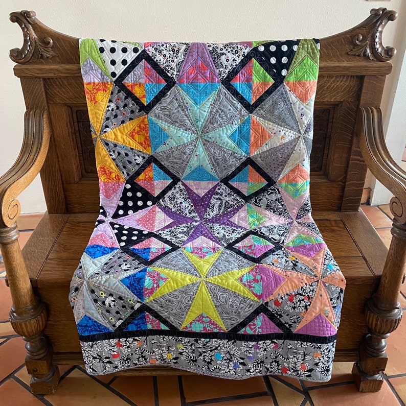 May include: A colorful patchwork quilt with star and diamond patterns, draped over a wooden bench. The quilt features various fabrics in shades of gray, black, yellow, orange, pink, purple, and blue. The quilt's border has a floral design.