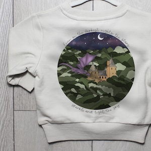 Puede incluir: Sudadera blanca con un diseño circular que muestra un dragón púrpura volando sobre un castillo en un bosque. El texto dice: "Even on the darkest nights the stars sparkle and light the sky."